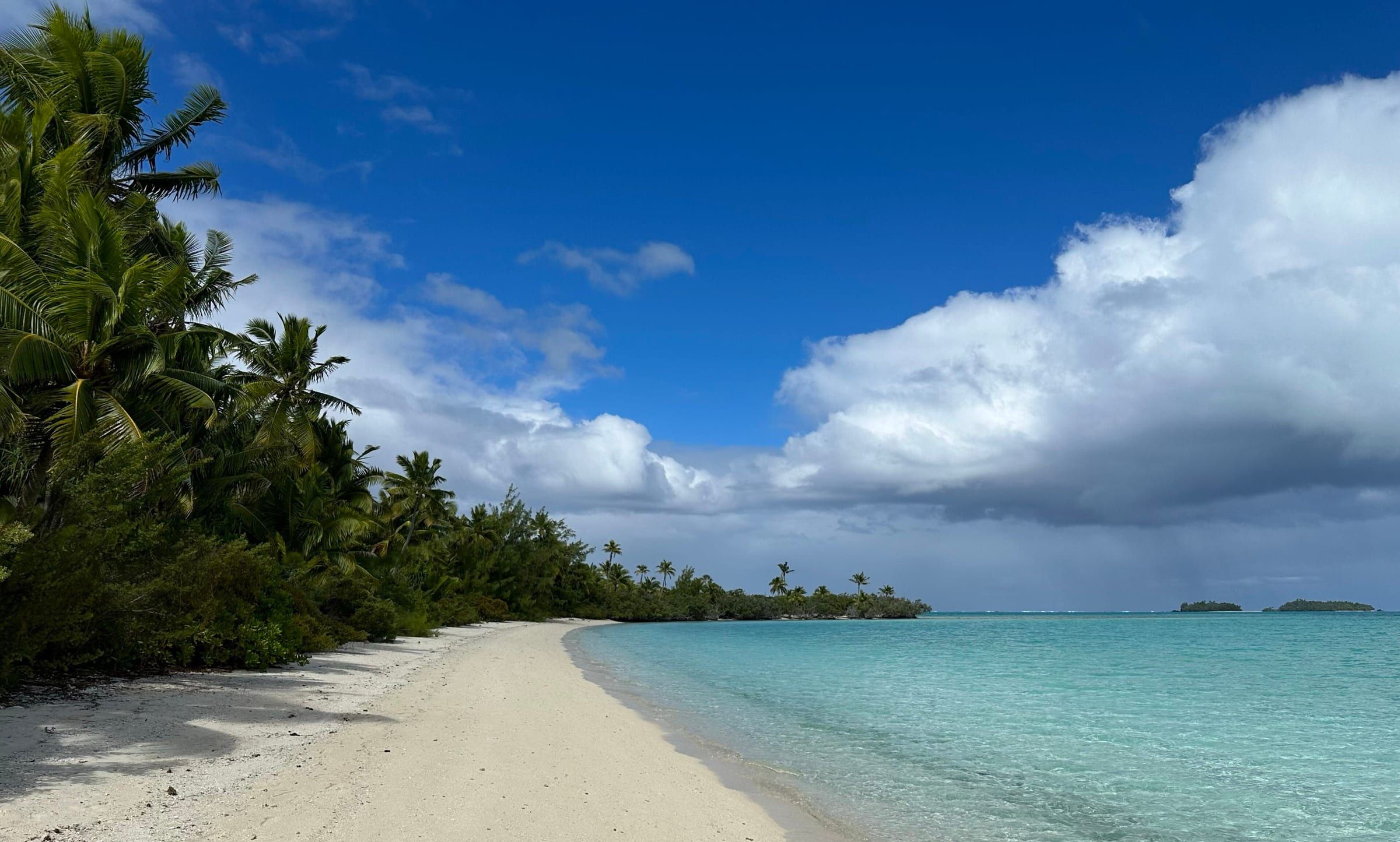 A curve of white sand and palms on Motu Akaiami, with clear turquoise water in Aitutaki lagoon and small islets on the horizon.