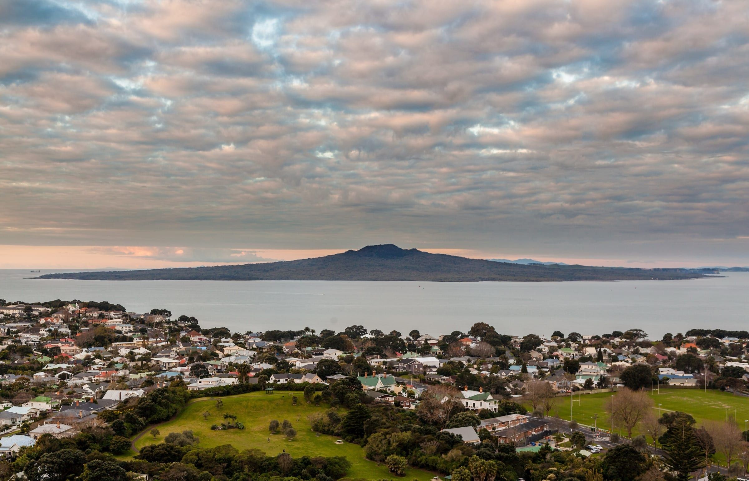 Rangitoto's low, even-sided volcanic cone stretched across the water beyond the rooftops and green reserves of Devonport, under a rippled pink-grey evening sky.