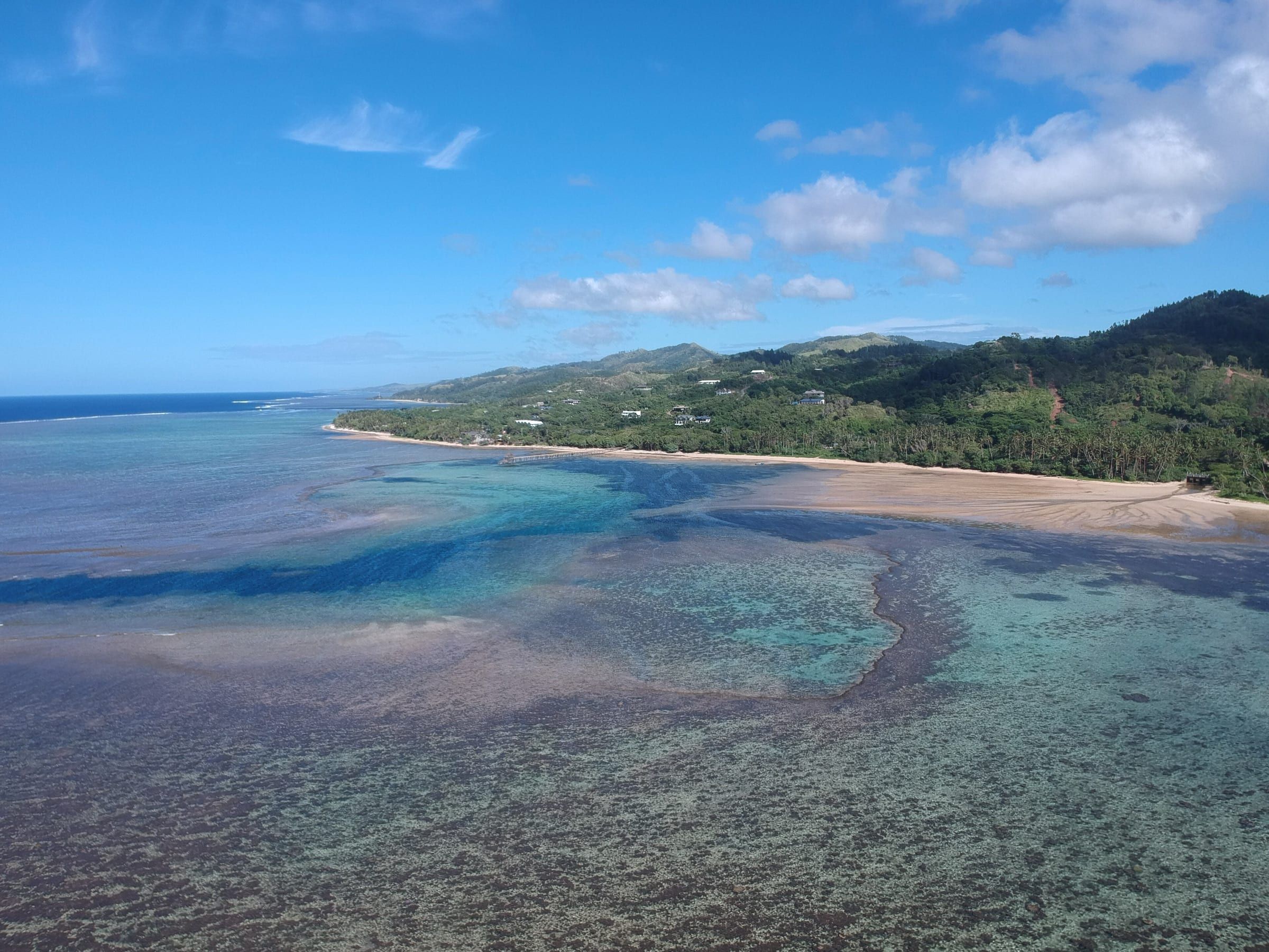 Aerial view of Viti Levu's Coral Coast: a wide reef flat and deep blue lagoon channel meet a sandy beach, a long jetty and forested hills dotted with houses.