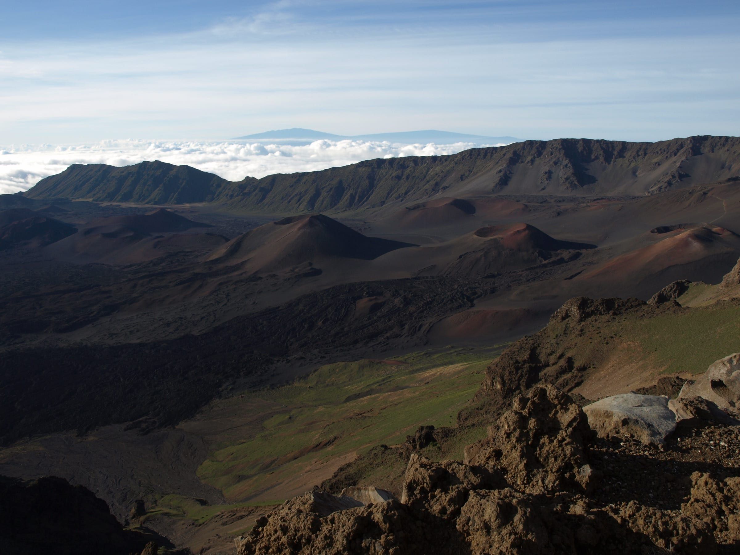 Rust-red and charcoal cinder cones scatter the floor of Haleakalā's summit crater on Maui, ringed by steep walls, with a sea of cloud and the Big Island's volcanoes beyond.