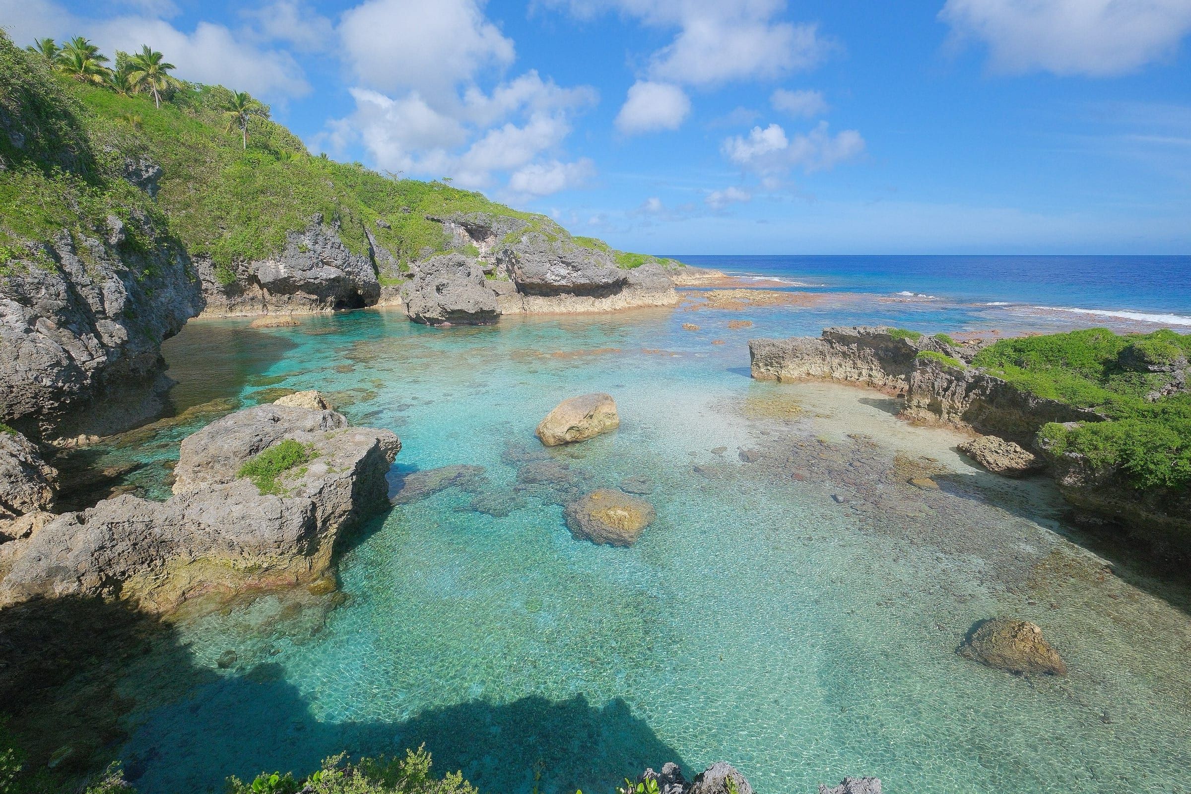 Limu Pools on Niue: a sheltered inlet of glassy turquoise water between grey limestone rocks and green slopes, opening onto the reef and deep-blue sea.