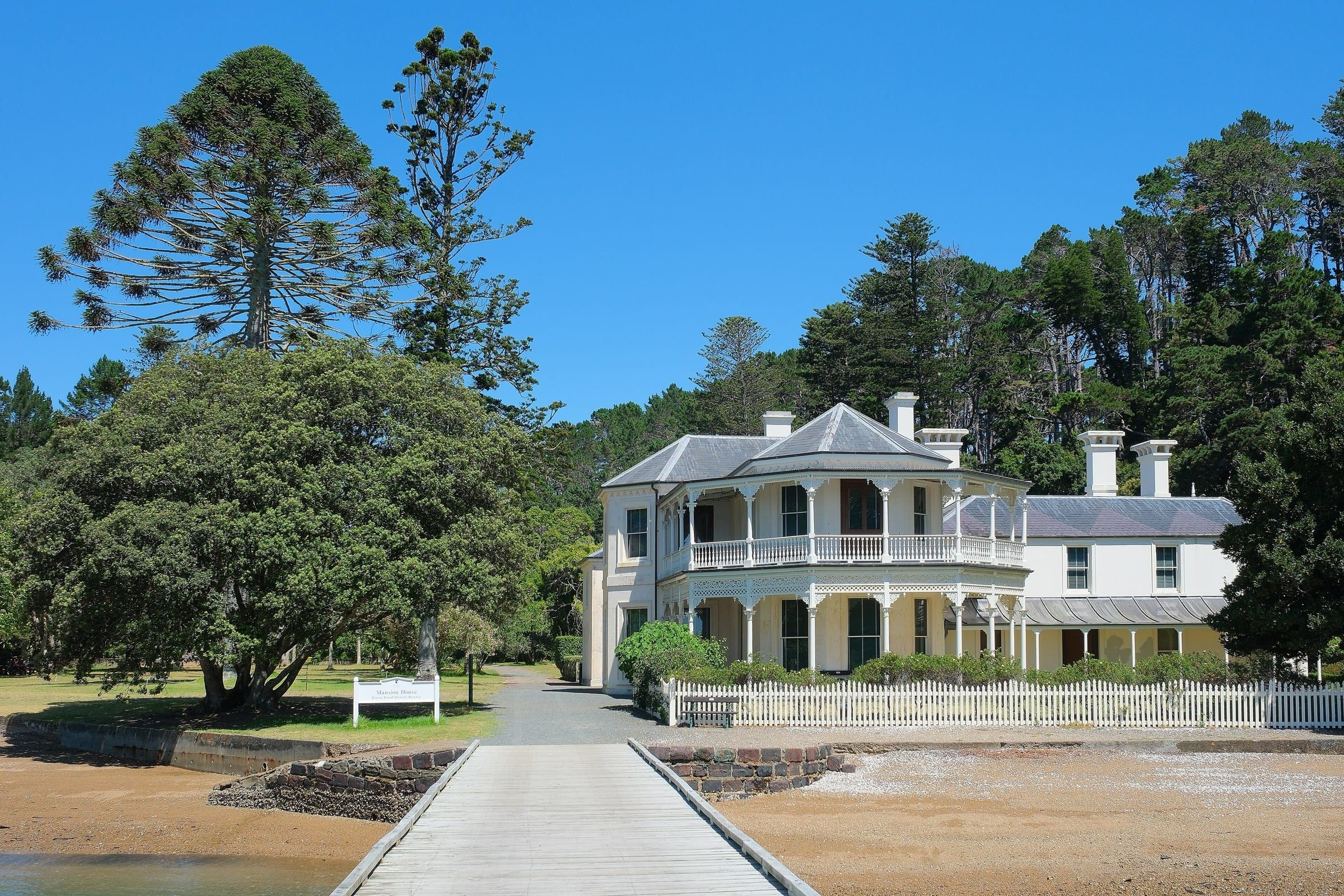 Mansion House on Kawau Island, a white two-storey Victorian house with an ornate verandah and picket fence, seen from the jetty across a sandy beach, under tall trees.