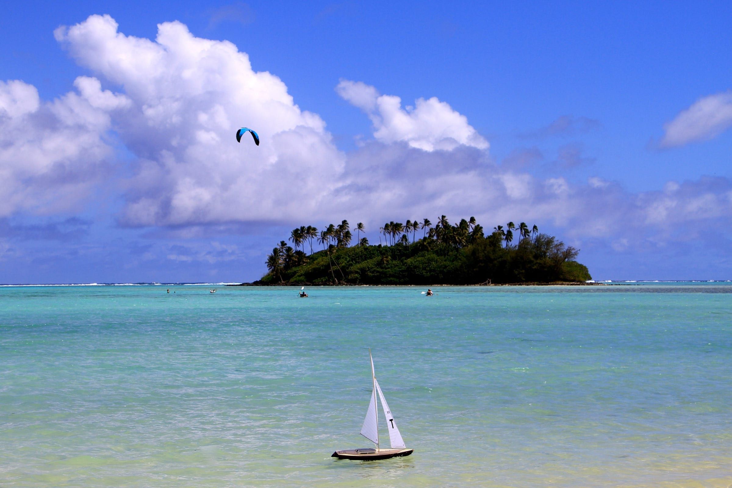 Clear turquoise shallows of Muri Lagoon on Rarotonga, looking out to the palm-topped islet of Taakoka, with a kite in the sky and a model yacht in the foreground.