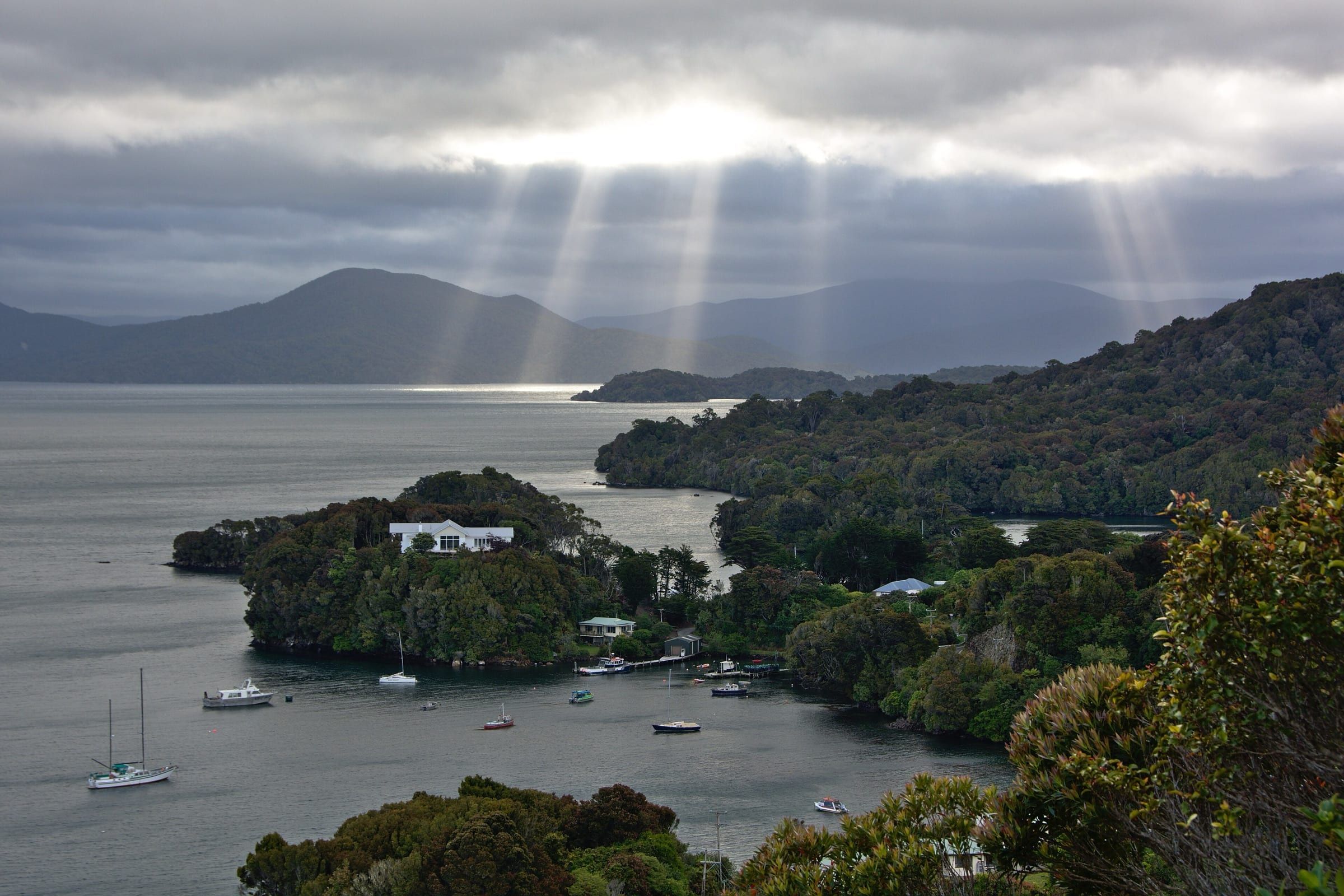 Shafts of sunlight break through heavy cloud over Paterson Inlet, lighting the water beyond a bush-covered headland and yachts moored in a sheltered bay below.