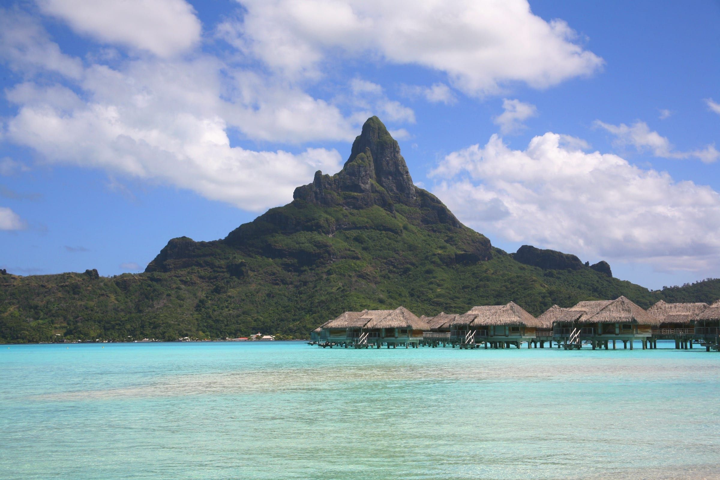 Mount Otemanu's sheer basalt peak rises above a pale turquoise lagoon, with a row of thatched overwater bungalows on stilts to the right.