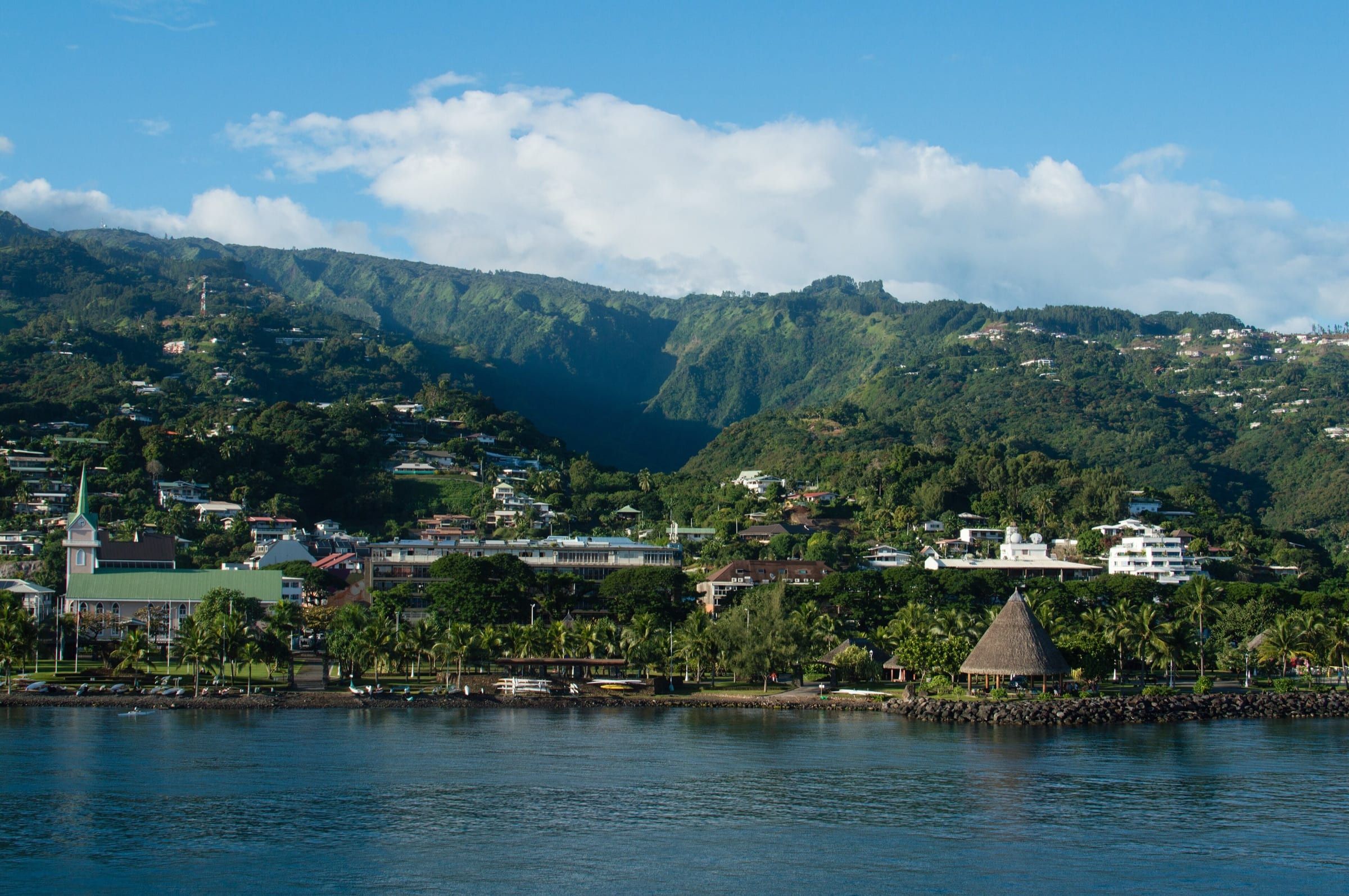 Papeete's waterfront at Pāʻōfaʻi on a sunny morning: palms, a steepled church and a thatched pavilion along the shore, with steep green valleys rising behind.