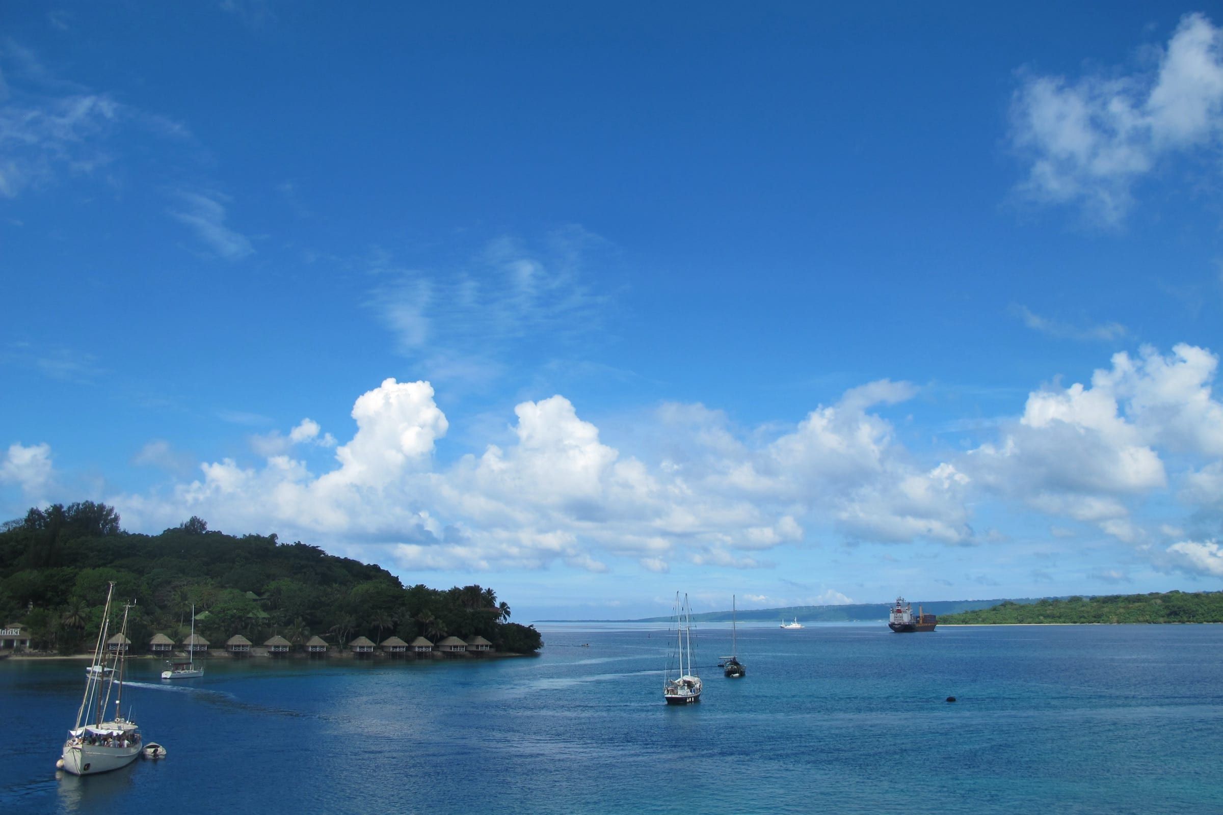 Yachts ride at anchor in Port Vila's blue harbour beside Iririki Island, whose thatched waterfront bungalows sit beneath forested slopes under a bright sky.