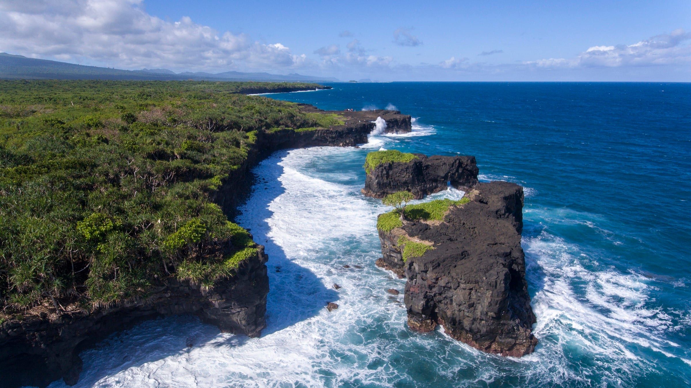 Aerial view of Upolu's south coast in O le Pupu Puʻe National Park: black lava cliffs and sea stacks ringed by white surf, dense green coastal forest and hills on the horizon.