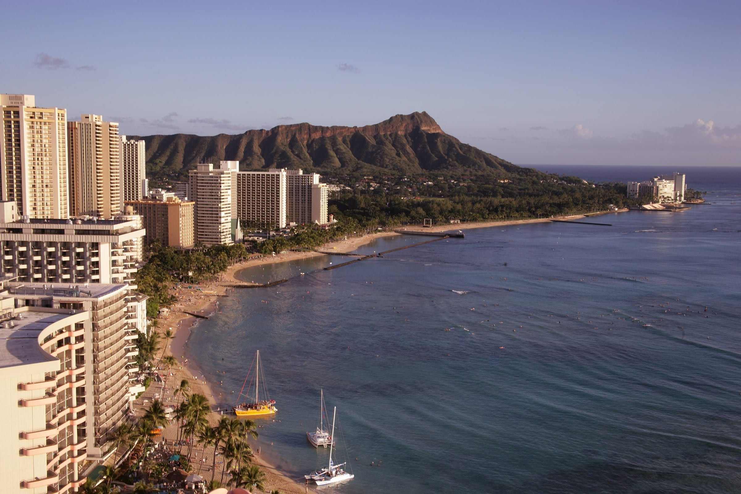 Late-afternoon light on Waikīkī: high-rise hotels and palms line the curving beach, sailboats sit offshore and the crater ridge of Diamond Head rises behind.