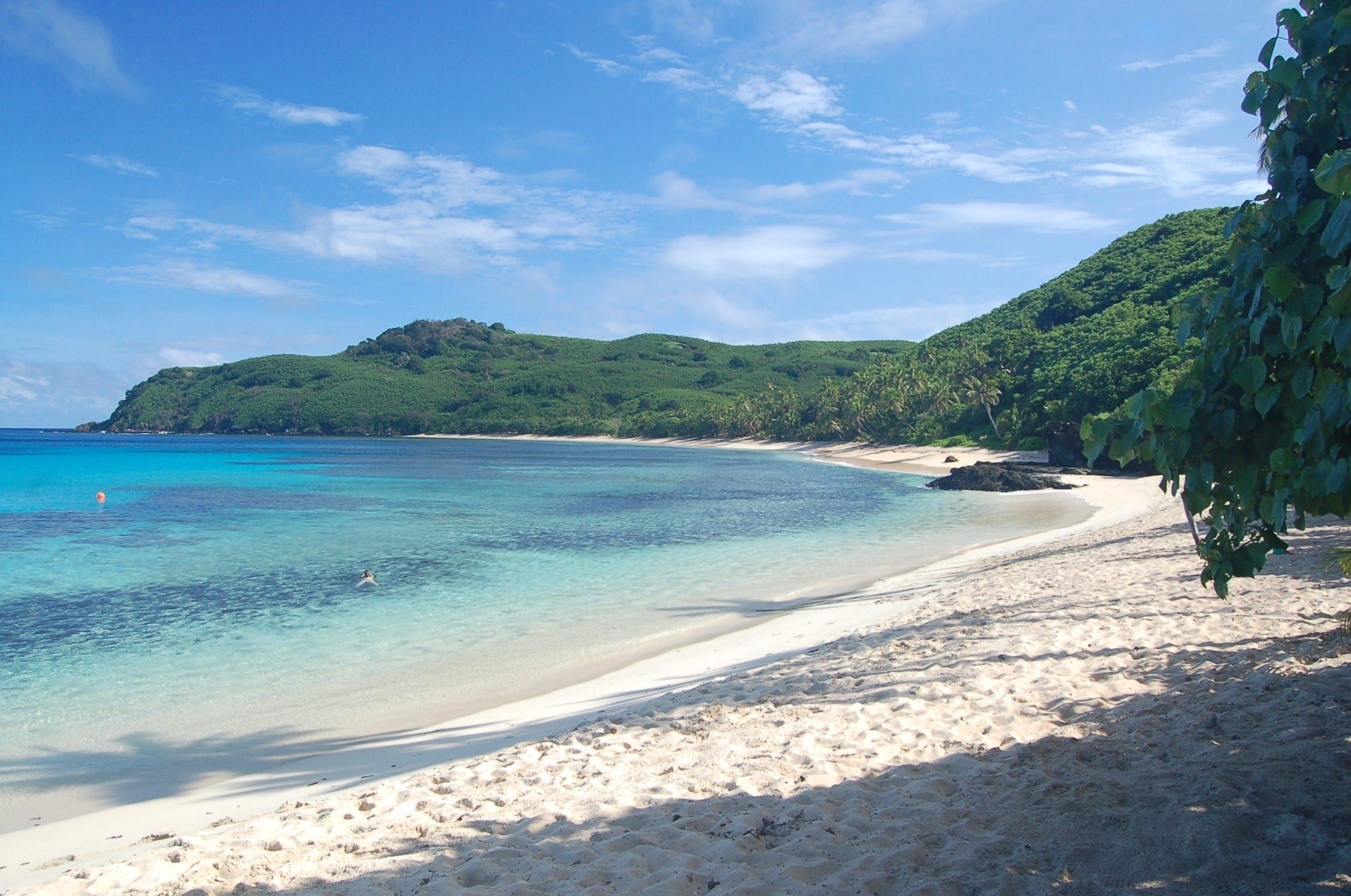 A curving white-sand beach on Waya, in the southern Yasawas, below a green forested hillside, with reef patches in the shallows and a lone swimmer.
